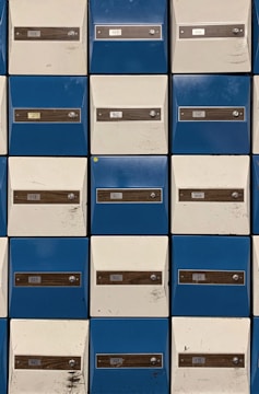 Rows of metal mailboxes with alternating blue and white doors, each labeled with a number and equipped with a lock. Some doors show marks and scratches.