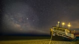 A fishing boat is stationed on a sandy beach under a starry night sky. The Milky Way galaxy is prominently visible, adding a mystical aura. The boat, named 'Roger Venture,' is illuminated by bright yellow lights that cast a warm glow on the surrounding area.