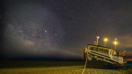 A fishing boat is stationed on a sandy beach under a starry night sky. The Milky Way galaxy is prominently visible, adding a mystical aura. The boat, named 'Roger Venture,' is illuminated by bright yellow lights that cast a warm glow on the surrounding area.