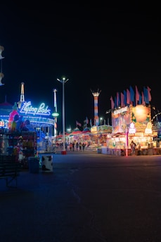 Brightly lit carnival at night featuring colorful rides and games. Neon signs and string lights illuminate the area, with various booths displaying snacks and prizes. A few people are walking along the midway, enjoying the festive atmosphere.