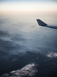 Aerial view of a landscape with an airplane's wing in the foreground. The scenery below shows densely packed clouds and a mountain range partially obscured by cloud cover. The sky transitions from a soft gradient of blues to pale tones near the horizon.