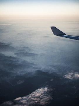 Aerial view of a landscape with an airplane's wing in the foreground. The scenery below shows densely packed clouds and a mountain range partially obscured by cloud cover. The sky transitions from a soft gradient of blues to pale tones near the horizon.