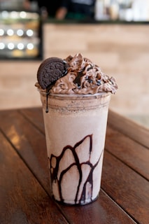 A tall glass filled with a creamy, chocolate-flavored beverage, topped with generous swirls of chocolate whipped cream and drizzled with chocolate syrup. An Oreo cookie is placed as a garnish on top of the whipped cream. The glass has a decorative pattern of chocolate syrup along the inside. The drink is placed on a wooden table, with a blurred background indicating a cafe setting.