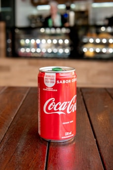A classic Coca Cola can resting on a wooden table with a bright, sunny outdoor café scene in the background.