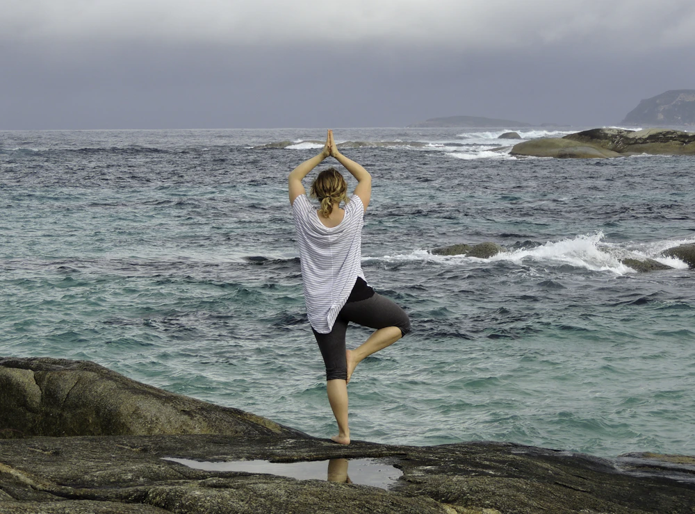 A woman in tree pose on sea rocks under a grey sky