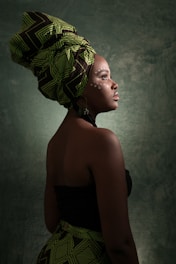 A person is dressed in traditional attire with a patterned headwrap and visible side profile. The skin features painted dots, enhancing the cultural theme. The background is textured and dark, highlighting the subject.