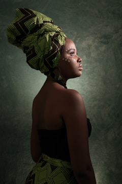 A person is dressed in traditional attire with a patterned headwrap and visible side profile. The skin features painted dots, enhancing the cultural theme. The background is textured and dark, highlighting the subject.