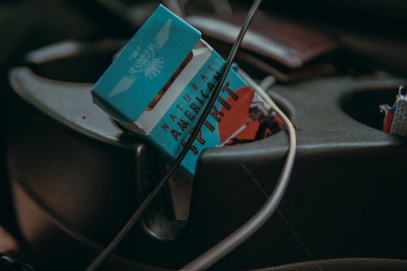 A pack of Natural American Spirit cigarettes is positioned in the center console of a car. The pack is open and partially visible. Various cables, possibly charging or audio wires, are tangled nearby, adding to a cluttered appearance.