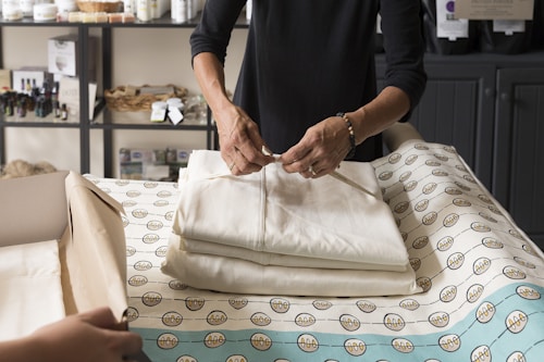 A person wrapping a folded garment in a retail or workshop setting. The table is covered with fabric that has a circular pattern, and a cardboard box is visible in the lower left corner. Shelves in the background hold various containers and baskets, suggesting a cozy and organized environment.