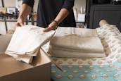 Hands are arranging neatly folded clothes on a table covered with patterned fabric. The scene appears to be in a room with shelves holding various bottles and boxes in the background.