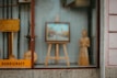 A shop window display contains a variety of handcrafted wooden items, including an easel with a framed picture, a standing wooden figure, and some other wooden objects. The word 'handicraft' is visible on a sign in the bottom left corner. The display conveys an artistic and artisanal vibe, with reflections on the glass slightly blurring the view.