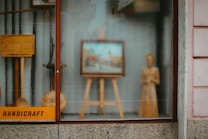 A shop window display contains a variety of handcrafted wooden items, including an easel with a framed picture, a standing wooden figure, and some other wooden objects. The word 'handicraft' is visible on a sign in the bottom left corner. The display conveys an artistic and artisanal vibe, with reflections on the glass slightly blurring the view.