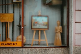A shop window display contains a variety of handcrafted wooden items, including an easel with a framed picture, a standing wooden figure, and some other wooden objects. The word 'handicraft' is visible on a sign in the bottom left corner. The display conveys an artistic and artisanal vibe, with reflections on the glass slightly blurring the view.