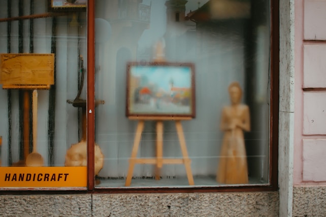 A shop window display contains a variety of handcrafted wooden items, including an easel with a framed picture, a standing wooden figure, and some other wooden objects. The word 'handicraft' is visible on a sign in the bottom left corner. The display conveys an artistic and artisanal vibe, with reflections on the glass slightly blurring the view.