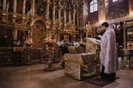 An ornately decorated interior with golden religious icons and motifs fills the space. A person in a decorative robe stands reading a book at a covered altar, surrounded by candles and elaborate gold detailing. The background features intricate paintings and carvings, with a somber and reverent atmosphere.