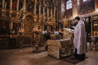 An ornately decorated interior with golden religious icons and motifs fills the space. A person in a decorative robe stands reading a book at a covered altar, surrounded by candles and elaborate gold detailing. The background features intricate paintings and carvings, with a somber and reverent atmosphere.