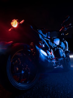 A motorcycle is illuminated by blue and red lighting, creating a dramatic contrast against the dark background. The focus is on the rear and side of the bike, highlighting its sleek design and intricate details. The red light casts a striking glow, enhancing the futuristic and edgy appearance of the vehicle.