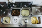 A buffet setup featuring multiple stainless steel containers, each containing different types of food including rice, roasted poultry, creamy soup, and French fries. Several of the containers have lids, and there are serving utensils visible, indicating a self-service food station.