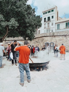 A group of people gathered around a large outdoor cooking pot, which is steaming heavily. It looks like a public event or gathering in a square, with people watching the cooking process. The setting includes a mature tree and older stone architecture with multi-story buildings in the background.