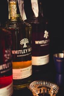 A close-up view of several bottles of Whitley Neill gin, prominently featuring a quince gin in the center. The label includes a detailed design of a tree and text about the gin. Around the bottles, a metal cocktail strainer and a jigger are visible, suggesting a bar setting.