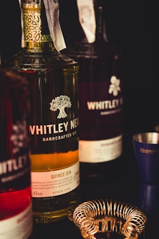 A close-up view of several bottles of Whitley Neill gin, prominently featuring a quince gin in the center. The label includes a detailed design of a tree and text about the gin. Around the bottles, a metal cocktail strainer and a jigger are visible, suggesting a bar setting.