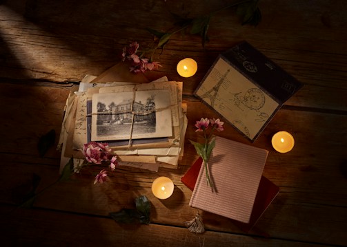 A wooden table is adorned with a collection of vintage letters, an old black and white photograph, and an assortment of pink flowers scattered among them. Three small lit candles provide a warm, ambient glow, casting soft shadows across the surface. An elegant box and a decorative notebook are also present, creating a nostalgic and intimate atmosphere.