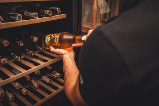 A person holding a bottle of peach-flavored wine or spirit, selecting it from a well-organized wooden wine rack containing multiple bottles stored horizontally. The scene suggests an indoor setting with warm lighting, emphasizing the amber color of the bottle.