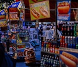 A bustling street scene featuring several hot dog stands displaying colorful signs and a variety of beverages. People can be seen in the background, walking through an urban environment. The stalls prominently advertise chili cheese dogs alongside other snacks, with pretzels visible in the foreground.