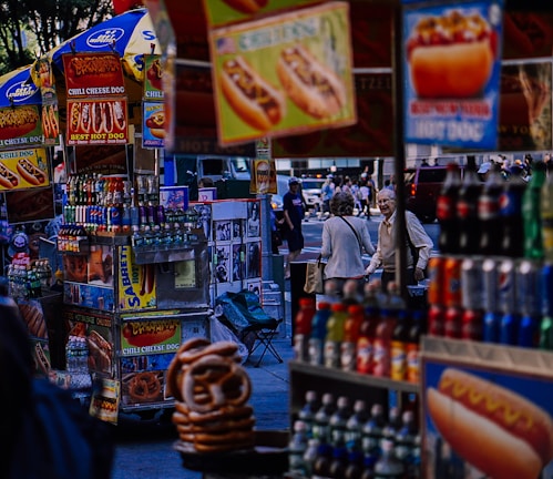 A bustling street scene featuring several hot dog stands displaying colorful signs and a variety of beverages. People can be seen in the background, walking through an urban environment. The stalls prominently advertise chili cheese dogs alongside other snacks, with pretzels visible in the foreground.