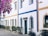 Colorful house with white walls and blue and yellow accents. The facade features arched windows and doors, surrounded by potted greenery. Vibrant purple flowers hang above, adding a lively touch.