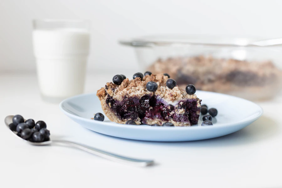 A slice of blueberry pie on a plate with fresh blueberries and a glass of milk