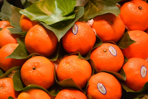 A collection of fresh, ripe oranges with vibrant orange peels, nestled among green leaves. Some oranges have small stickers on them, likely indicating their brand or origin. The natural texture and shine of the fruit are clearly visible.