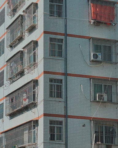 A residential building's facade with barred windows, each covered with metal grilles. Air conditioning units and some items like clothing or fabric can be observed hanging outside the windows. The building is tiled with a grid pattern, incorporating subtle colored stripes.
