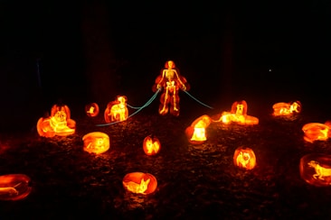 The image features a group of intricately carved pumpkins illuminated from within, showcasing various designs. At the center is a large skeleton figure holding what appear to be leashes that are connected to animal-shaped pumpkin carvings around it. The scene is set against a dark background, highlighting the glowing orange of the pumpkins.