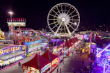 A vibrant carnival scene at night featuring a large Ferris wheel illuminated against the dark sky. Numerous stalls are lined up with colorful lights and decorations, including games and food vendors. Crowds of people walk through the area, enjoying the festive atmosphere. Bright signage and banners add to the lively ambiance.