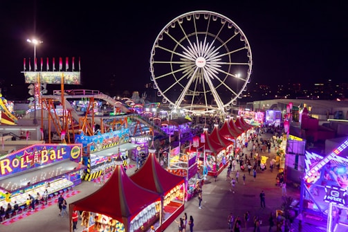 A vibrant carnival scene at night featuring a large Ferris wheel illuminated against the dark sky. Numerous stalls are lined up with colorful lights and decorations, including games and food vendors. Crowds of people walk through the area, enjoying the festive atmosphere. Bright signage and banners add to the lively ambiance.