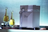A luxurious gift box adorned with a ribbon featuring the Dior brand name is placed alongside two elegant bottles of perfume on a reflective surface. The background exhibits a soft gradient of colors, enhancing the sophisticated and exquisite presentation.