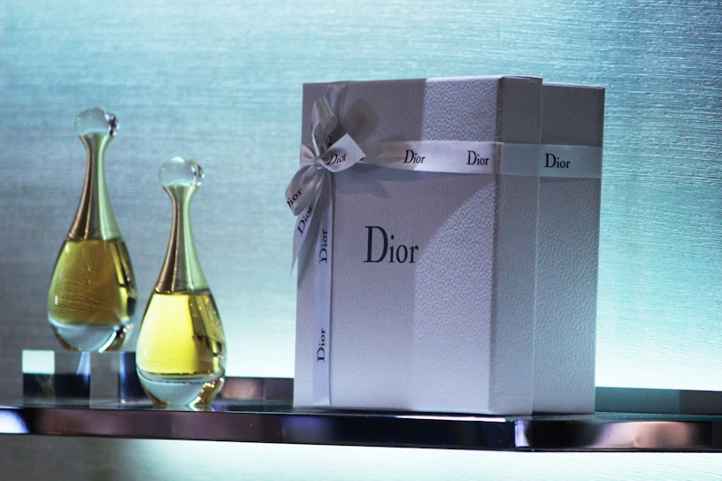 A luxurious gift box adorned with a ribbon featuring the Dior brand name is placed alongside two elegant bottles of perfume on a reflective surface. The background exhibits a soft gradient of colors, enhancing the sophisticated and exquisite presentation.