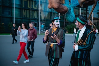 Two street performers dressed in colorful costumes, with face paint and hats, engage with each other on a city street. One performer gestures while the other listens. Passersby, including a woman using a smartphone, walk behind them near a modern glass building.