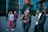 Two street performers dressed in colorful costumes, with face paint and hats, engage with each other on a city street. One performer gestures while the other listens. Passersby, including a woman using a smartphone, walk behind them near a modern glass building.