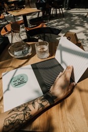 A tattooed arm resting on a wooden table, with a hand holding open a notebook. On the table are a glass carafe and a water glass. In the background, there are wooden chairs and tables in an open, sunlit space.