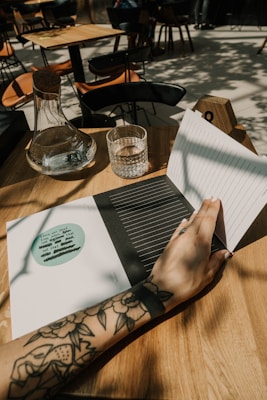 A tattooed arm resting on a wooden table, with a hand holding open a notebook. On the table are a glass carafe and a water glass. In the background, there are wooden chairs and tables in an open, sunlit space.
