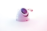 A white surveillance camera with a dome shape is positioned on a plain white background, reflecting a minimalistic aesthetic. The camera lens is prominently displayed in the center, surrounded by several reflective elements. The cable connected to the camera is visible in the background, adding to the industrial feel of the object.