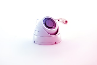 A white surveillance camera with a dome shape is positioned on a plain white background, reflecting a minimalistic aesthetic. The camera lens is prominently displayed in the center, surrounded by several reflective elements. The cable connected to the camera is visible in the background, adding to the industrial feel of the object.
