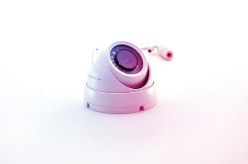 A white surveillance camera with a dome shape is positioned on a plain white background, reflecting a minimalistic aesthetic. The camera lens is prominently displayed in the center, surrounded by several reflective elements. The cable connected to the camera is visible in the background, adding to the industrial feel of the object.