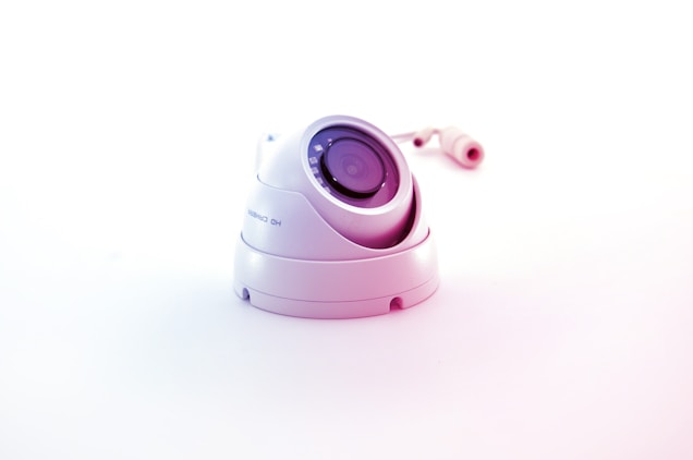 A white surveillance camera with a dome shape is positioned on a plain white background, reflecting a minimalistic aesthetic. The camera lens is prominently displayed in the center, surrounded by several reflective elements. The cable connected to the camera is visible in the background, adding to the industrial feel of the object.