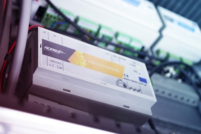A close-up of an electrical device with various labels and indicators, surrounded by wires and connectors. The device appears to be mounted on a panel or within an enclosure.