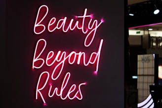 Neon pink script reads 'Beauty Beyond Rules' against a dark background with a partially visible sign in the background.