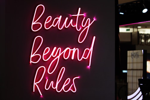 Neon pink script reads 'Beauty Beyond Rules' against a dark background with a partially visible sign in the background.