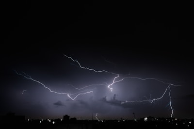 A dramatic night sky is illuminated by multiple streaks of lightning, creating a web-like pattern across the dark clouds. The ground below is silhouetted, adding to the intensity and scale of the storm.